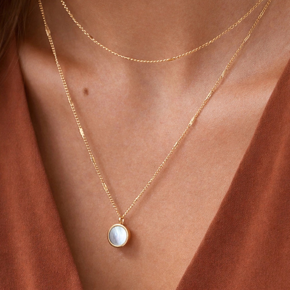 Layered gold chain necklaces with a mother-of-pearl pendant worn over rust silk